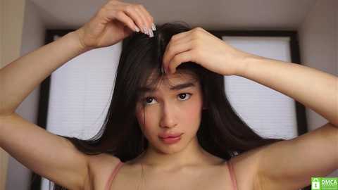 Video of a young Asian woman with long black hair, fair skin, and a slender build, combing her hair in a room with white blinds. She wears a pink spaghetti-strap top.