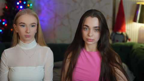 Media: Video of two young women with long hair, one in a sheer white top and the other in a bright pink dress, seated indoors, Christmas lights in the background.