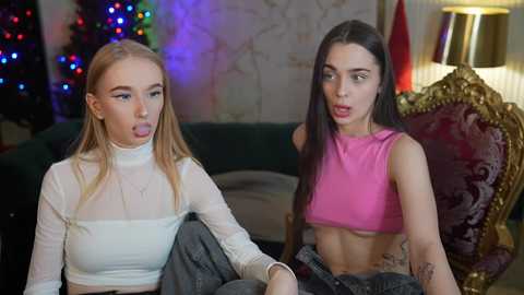 Media: Video of two young women in festive attire: one blonde in a sheer top, the other brunette in a pink crop top. Background features a Christmas tree, a gold chair, and a lamp.