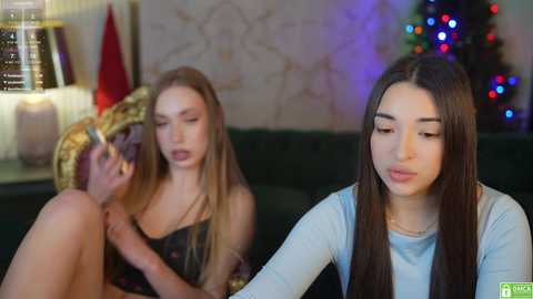 Media: Video of a young woman with long dark hair, wearing a light blue top, pouting while sitting on a dark couch. Background features a blonde woman in a patterned dress, holding a cowboy hat, and a decorated Christmas tree.