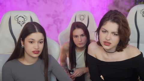 Media: Video of three young women with pale skin, dark hair, and neutral expressions sitting in gaming chairs against a purple-lit wall. They wear casual, off-the-shoulder black tops.