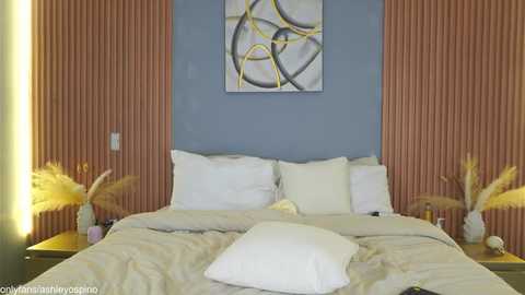 A video of a modern bedroom with a white bed, two white pillows, beige bedding, a blue accent wall, and abstract art; two side tables with potted plants and lamps.