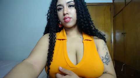 Media: Video of a light-skinned woman with curly black hair, wearing a tight orange top that accentuates her large breasts and tattoos. She stands indoors, looking at the camera with a slight pout.