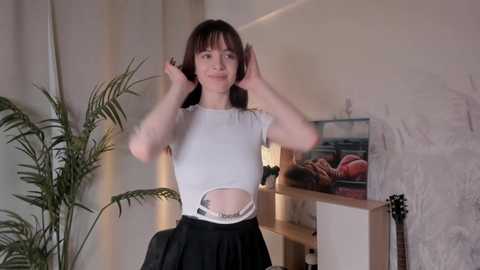 Media: Video of a young, slender, fair-skinned woman with shoulder-length brown hair, wearing a white crop top revealing her midriff and black pleated skirt. She stands in a cozy, dimly lit room with a plant, wall art, and a lamp.