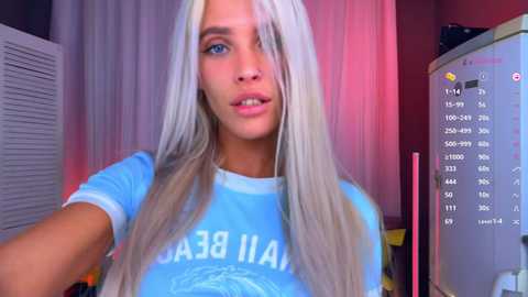 Video of a young woman with platinum blonde hair, fair skin, and blue eyes, wearing a light blue t-shirt. She stands in a dimly lit room with a white wall, pink curtains, and a computer monitor displaying a weather app.