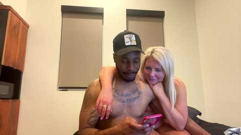 Media: A video captures a shirtless Black man with a tattooed chest and a blonde woman in a room. They're seated on a bed, the man holding a smartphone while the woman rests her head on his shoulder.