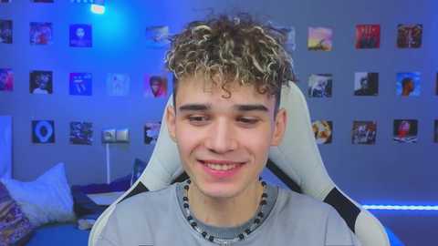 Video of a young man with curly, blonde hair, grinning, wearing a grey shirt, seated in a white gaming chair, in a brightly lit, blue-themed room with numerous framed posters on the wall.