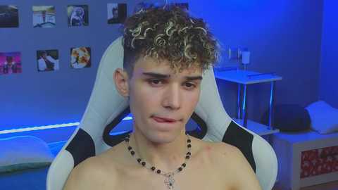 Media: Video of a shirtless young man with curly hair and a necklace, seated in a white gaming chair in a dimly lit room with blue lighting, photos on the wall, and a desk.