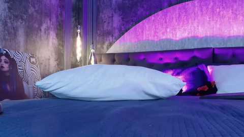 A video of a dimly lit, modern bedroom with a purple-hued headboard, two white pillows, and a striped pillow on a blue quilted bed. A person in black and red clothing is partially visible beneath the bed.