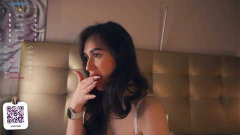 Media: Video of a young Asian woman with long black hair, wearing a white sleeveless top, and an Apple Watch, looking contemplatively with her finger to her lips. Background features a beige cushioned headboard and a poster with text.