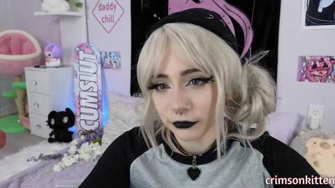 Video of a pale-skinned woman with platinum blonde hair in pigtails, black makeup, and a black and gray shirt, in a pink-themed bedroom. Background features a cat, plush toys, and pastel decor.