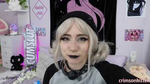 Video of a young woman with platinum blonde hair, wearing a black and grey hoodie, black lipstick, and a black beanie. Background includes pastel decor, plush toys, and framed art.