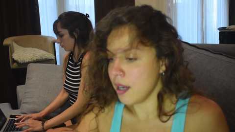 Video of two young women, one with curly hair in a blue top, and the other with straight hair in a striped shirt, sitting on a couch in a modern living room with beige curtains.