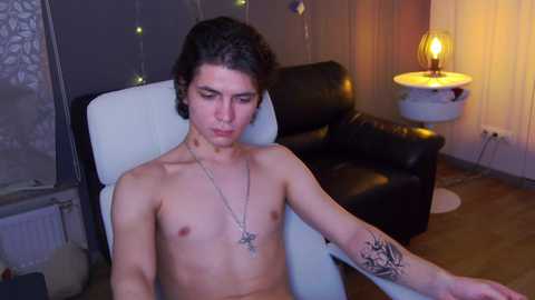 Video of a shirtless, slender young man with curly black hair, tattoos on his arm, sitting in a white chair, looking contemplative. Background includes a black leather couch, wall sconce, and string lights.