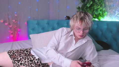 Media: Video of a woman with short blonde hair and glasses, wearing a white shirt and leopard print skirt, lying on a bed with a turquoise headboard, looking at a smartphone. Background features a green plant and multicolored fairy lights.