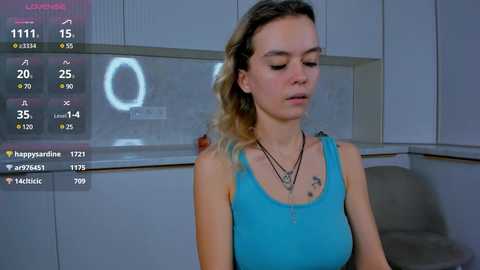 Media: Video of a Caucasian woman with wavy blonde hair, wearing a blue tank top, in a modern kitchen with white cabinets, a round ring light, and a gray chair.