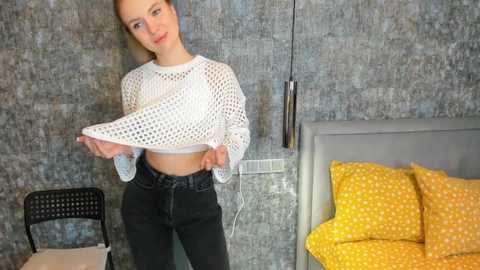 Media: Video of a fair-skinned, blonde woman in a white mesh crop top, black jeans, and yellow polka-dot pillow, posing in a modern, gray-walled bedroom with a black chair and a gray upholstered headboard.