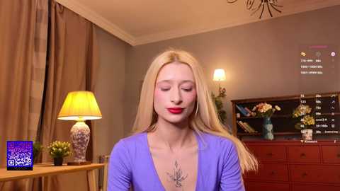 Media: Video of a fair-skinned, blonde woman with long hair, wearing a purple top, standing indoors. Background includes a lamp, a dresser, and a clock, with a \"TV Service\" logo in the bottom left corner.