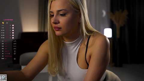 Video of a blonde Caucasian woman with fair skin, wearing a white tank top, looking at a laptop screen with social media notifications, in a dimly lit room with a wooden chair and dark curtains.