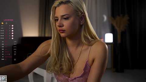 A video of a blonde woman in a pink lace bra, sitting indoors with a dimly lit background featuring a modern decor and a digital interface on the left.