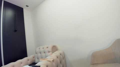 Media: A video of a minimalist bedroom with a beige tufted sofa against a white wall, a dark wooden door, and a small wooden stool. The room's simplicity emphasizes a clean, modern aesthetic.