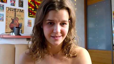 Video of a young woman with wavy brown hair, fair skin, and a gentle smile, wearing a gold necklace. Background features colorful posters, a guitar, and a wooden door.