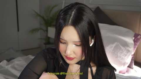 Video of an East Asian woman with long black hair, fair skin, and red lipstick, wearing a black sheer top, sitting on a bed with white pillows, looking down, in a dimly lit bedroom.