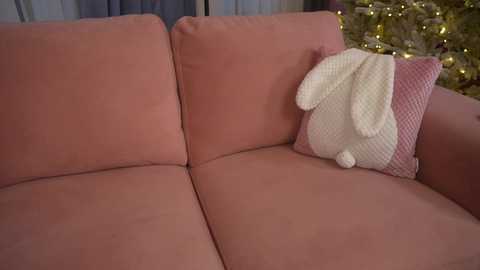 Video of a cozy living room with a soft pink couch featuring a white knitted rabbit pillow. The background includes a Christmas tree with golden ornaments and warm, soft lighting.