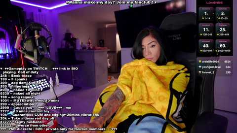 A screenshot from a Twitch stream shows a young woman with dark hair, wearing a yellow jacket, reclining on a black chair in a dimly lit room. The background includes a computer setup with various gaming equipment. Text chat with comments is visible.