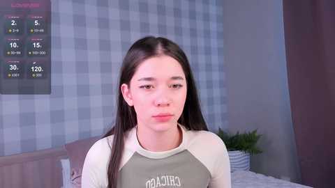 Video of a young Asian woman with long black hair, wearing a grey and white sweater, standing in a modern bedroom with checkered wallpaper and a potted plant.