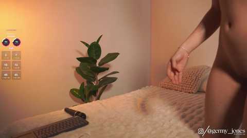 Video of a nude woman with light skin, standing on a beige carpeted room with a potted plant and a textured beige blanket on a bed.