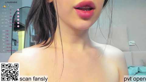 Video of an Asian woman with fair skin and full lips, partially visible due to the angle, wearing no top, standing in a modern room with a calendar and a lamp in the background.