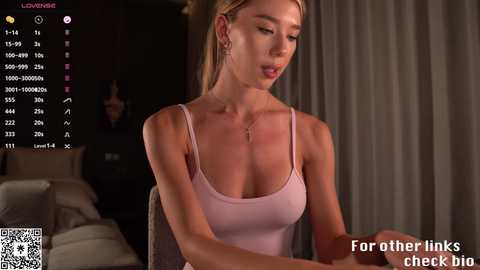 Video of a blonde woman with fair skin, wearing a pink tank top, seated at a desk, looking at a computer screen.