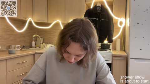 Video of a woman with shoulder-length brown hair in a light gray sweater, sitting in a modern kitchen with wooden cabinets, a hanging jacket, and glowing neon lights forming a shower shape, with text overlay: \"shower show is about to start.\