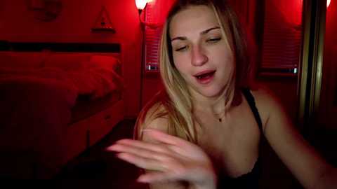 Media: Video of a blonde woman with fair skin, wearing a black bra, mouth open in apparent pleasure, in a dimly lit room with red lighting, a bed visible in the background.