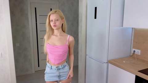 Media: Video of a slender, blonde Caucasian woman with fair skin, wearing a pink crop top and light blue shorts, standing in a modern, minimalist apartment with white walls and a refrigerator.