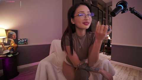 Media: Video of a slender Asian woman with long black hair, wearing glasses and a brown t-shirt, sitting on a chair in a dimly lit room, holding a microphone.