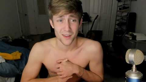 Media: Video of a shirtless young man with light skin and short brown hair, holding his chest in a bedroom with a blue bed, white door, and a microphone.