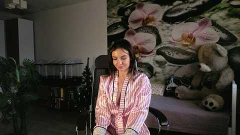 Media: Video of a young woman with long black hair, wearing a pink and white striped robe, sitting in a chair. Background features a floral painting and stuffed toys.