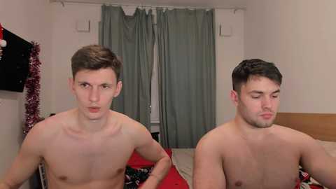 Media: Video of two shirtless young men with fair skin, dark hair, and lean physiques, seated side-by-side on a bed, in a minimalistic bedroom with green curtains and a festive red and white blanket.