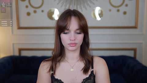 Media: Video of a young Caucasian woman with light skin, straight brown hair, and bangs, wearing a black lace bralette and gold necklace, seated on a dark blue velvet couch. Background features a white wall with gold circular patterns and a chandelier.