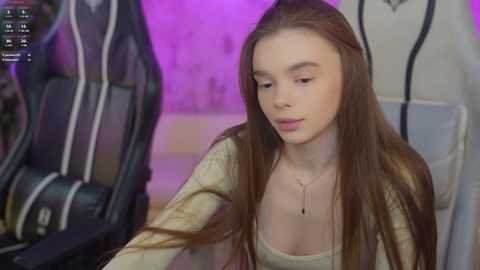 A video of a young Caucasian woman with long brown hair, wearing a beige sweater and necklace, sitting in front of a gaming chair, with purple lighting in the background.