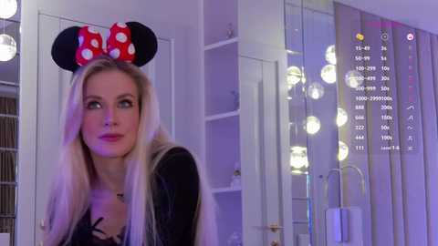 Video of a blonde woman with straight hair, wearing a red polka-dot Minnie Mouse headband, black dress, and makeup, standing in a room with white walls, mirrors, and glowing round lights.