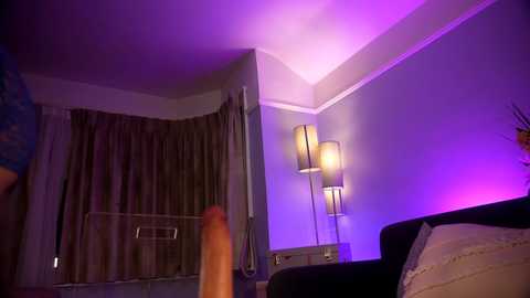 Video of a dimly-lit, modern bedroom with purple lighting. A large, erect penis is visible in the foreground, while a man in a blue shirt stands near a bed with beige sheets.