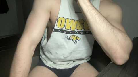 Media: Video of a slender, fair-skinned person wearing a white \"OWU\" tank top and black shorts, sitting with legs apart, left arm raised. Background is dimly lit, with a dark object in the lower left.