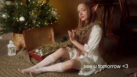 Media: Video of a young woman with pointed ears, wearing a white nightgown and knee-high socks, sitting on a beige rug, stringing tinsel around a red open box next to a decorated Christmas tree.