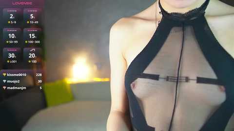 A video of a woman wearing a sheer black top that reveals her small breasts and nipples. The background shows a dimly lit bedroom with a bed and a glowing lamp.