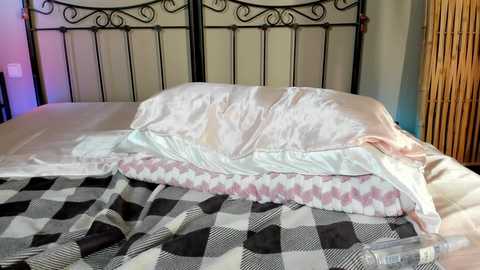 A video of an unmade bed with a black metal headboard, a white duvet, a light pink blanket, and a checkered black and white blanket. The room has bamboo blinds and a cozy, rustic atmosphere.