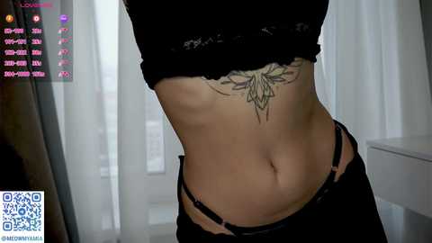 Video of a woman's midriff in a black lace bralette, revealing a detailed flower tattoo below her navel. Background shows a white curtain and a white dresser, with social media icons in the top left corner.