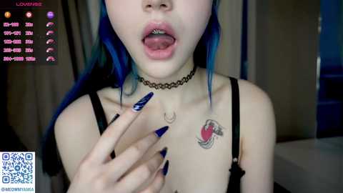 Video of a young woman with blue hair, wearing a black choker, black bra, and dark blue nail polish, licking her lips, in a dimly lit room.